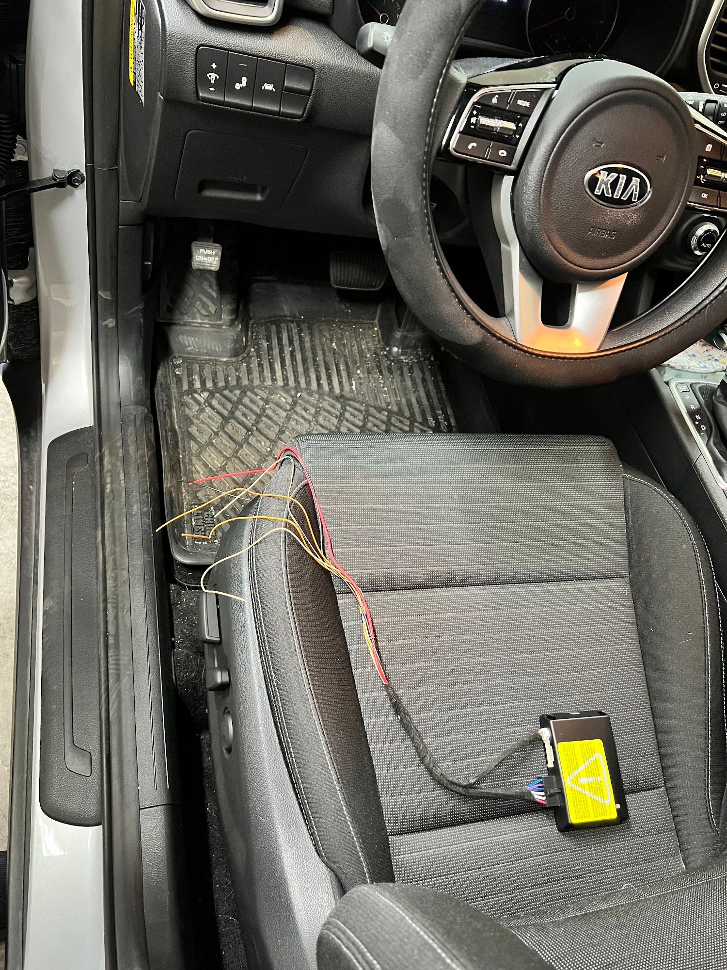 Interior view of a Kia car. A device with wires is placed on the driver's seat, likely related to vehicle modifications.