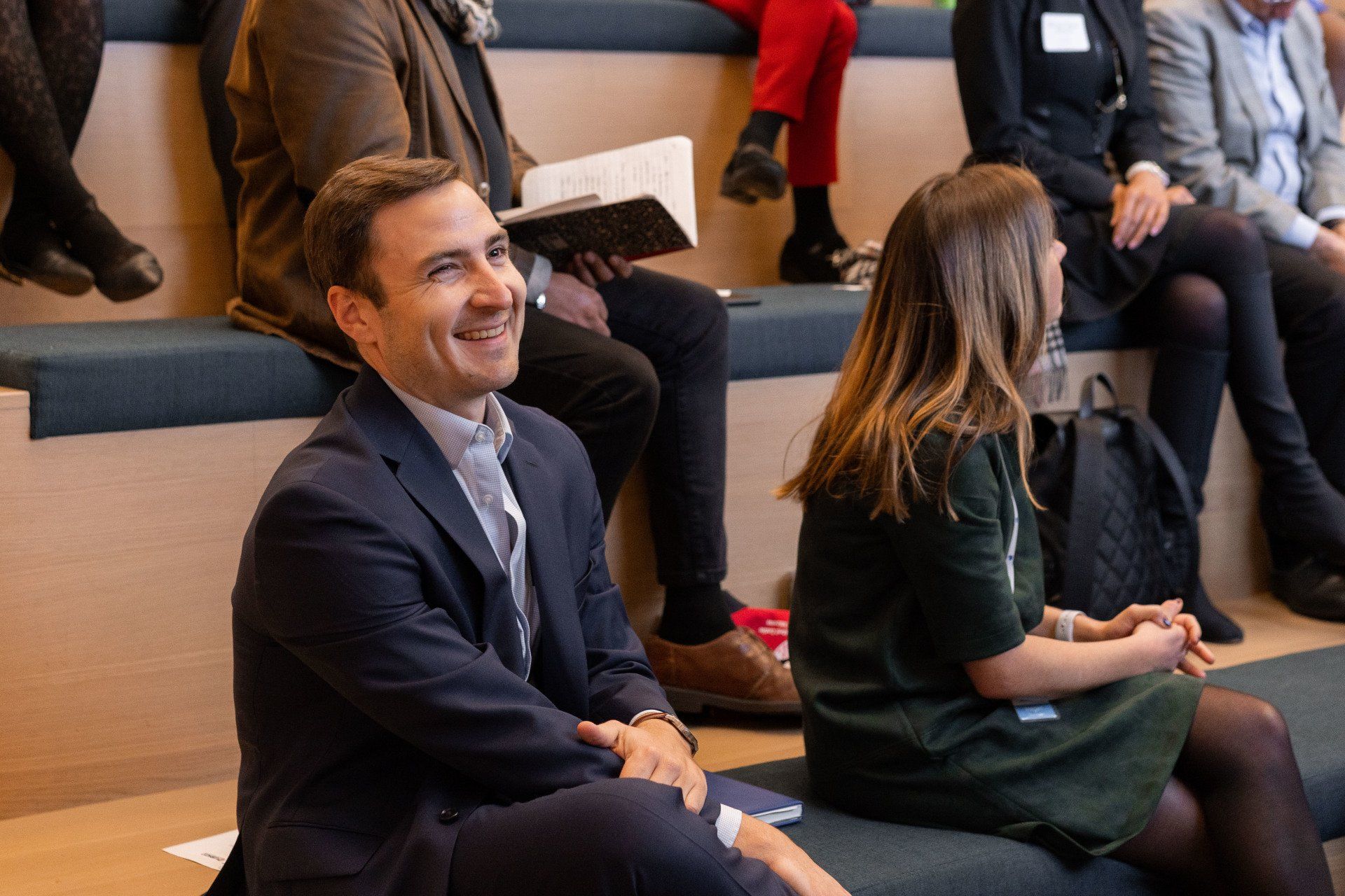 A man in a suit is smiling while sitting on a bench with other people.