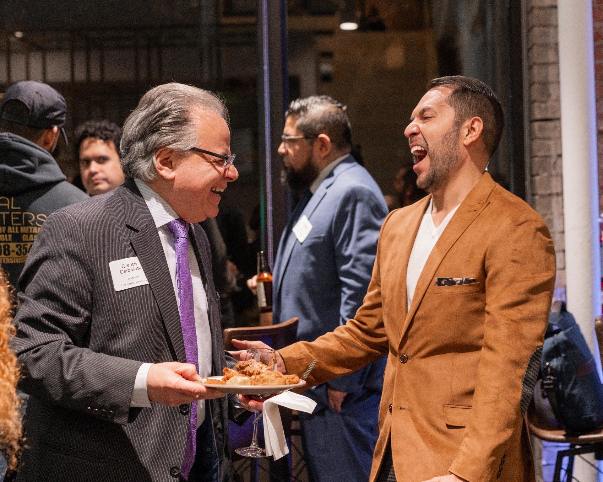 A man in a suit and tie is holding a plate of food and laughing with another man.