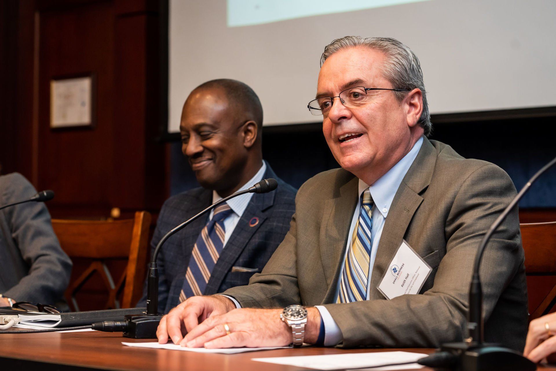 A man in a suit and tie is sitting at a table talking into a microphone.
