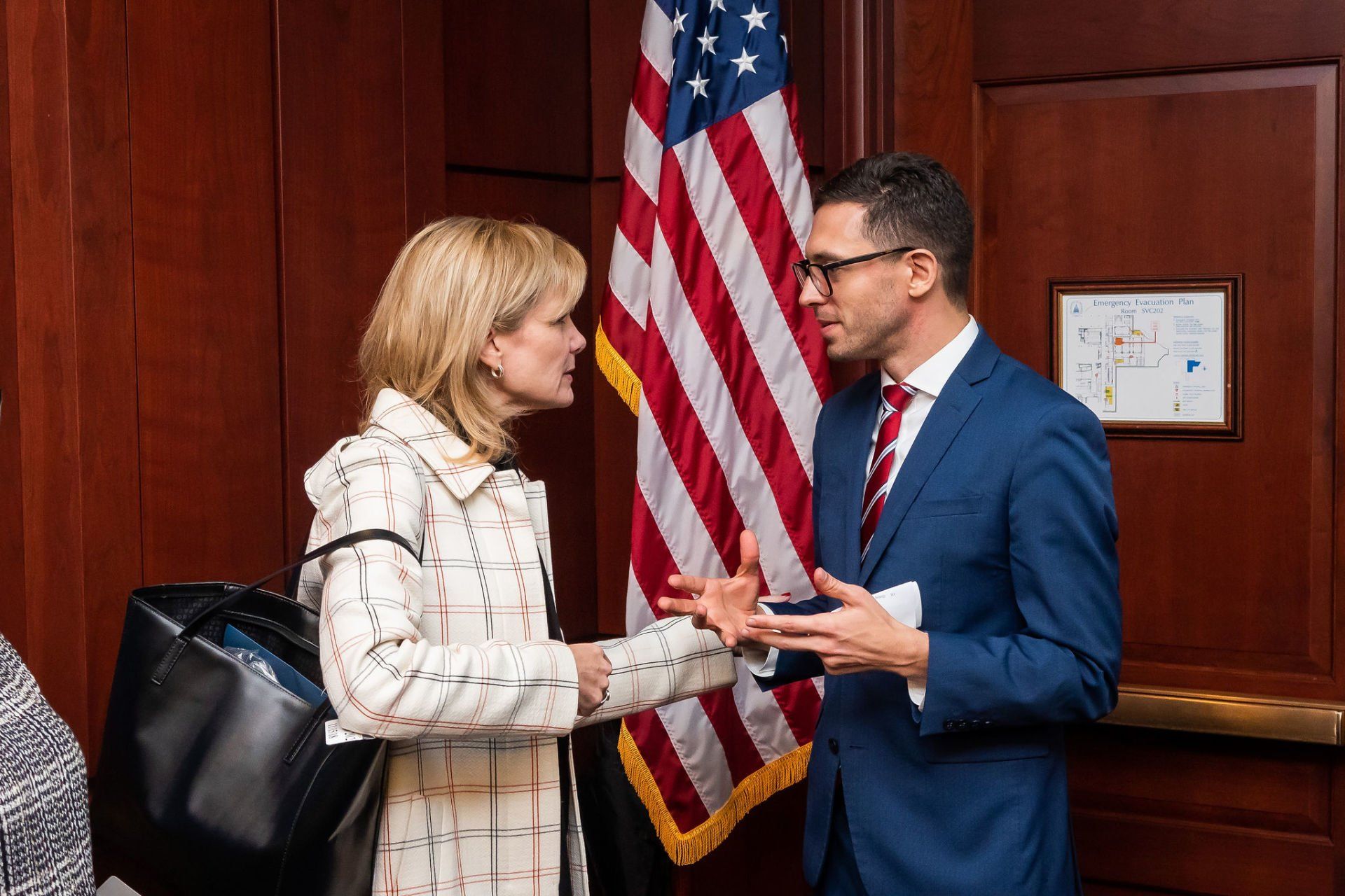 A man in a suit and tie is talking to a woman in front of an american flag.