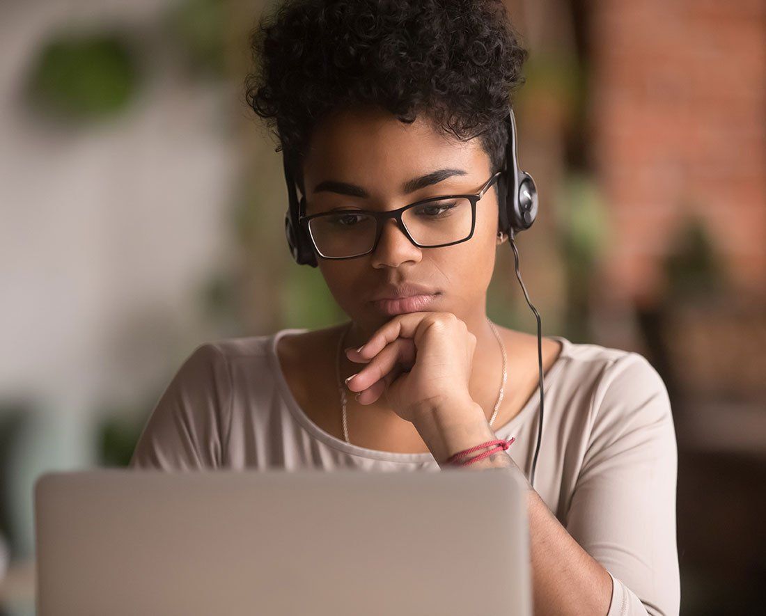 Young Black woman with glasses and headphones, working on a laptop, focused and contemplative.
