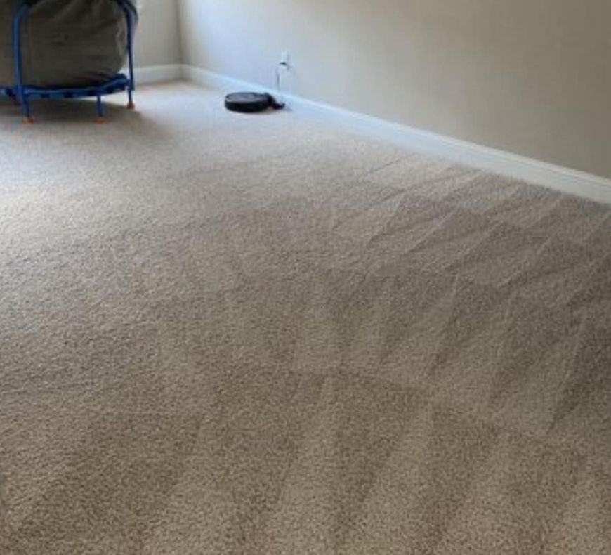 Empty beige carpeted room with a white wall, baseboard, and a few items near the far corner.