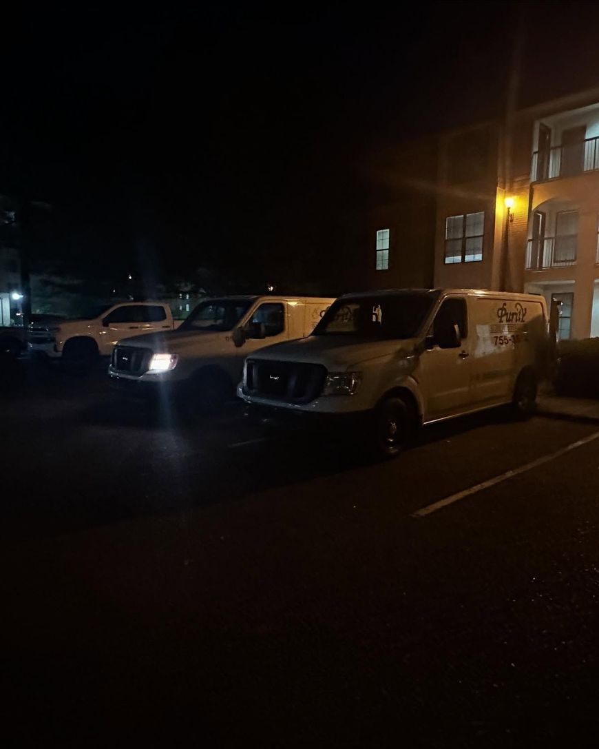 Two white vans parked at night beside a lit building, with headlights shining.