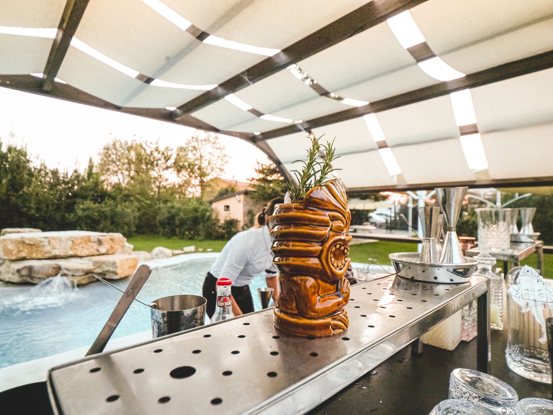 A brown tiki mug with a garnish sits on a stainless steel bar counter next to an outdoor swimming pool at sunset.