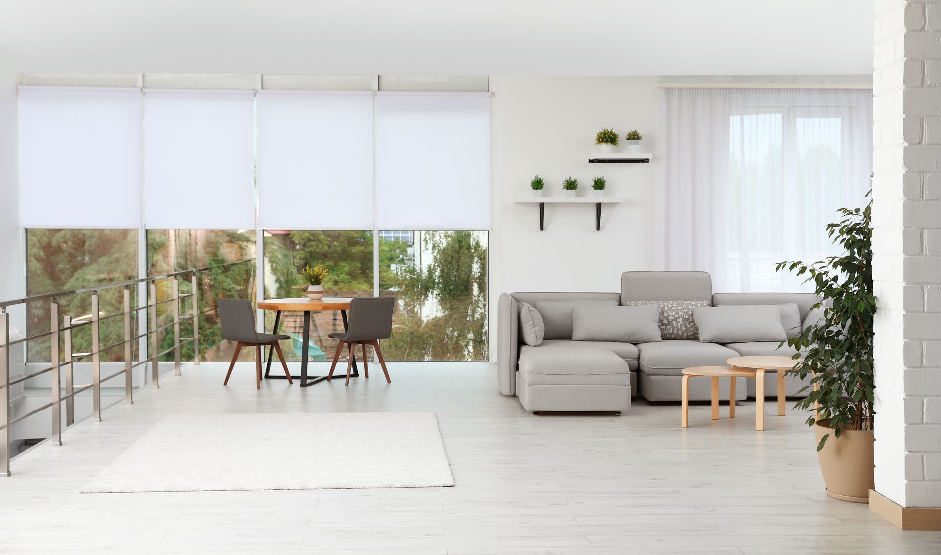 A bright, modern living room featuring white roller blinds, a gray sectional sofa, a small dining set, and potted plants.