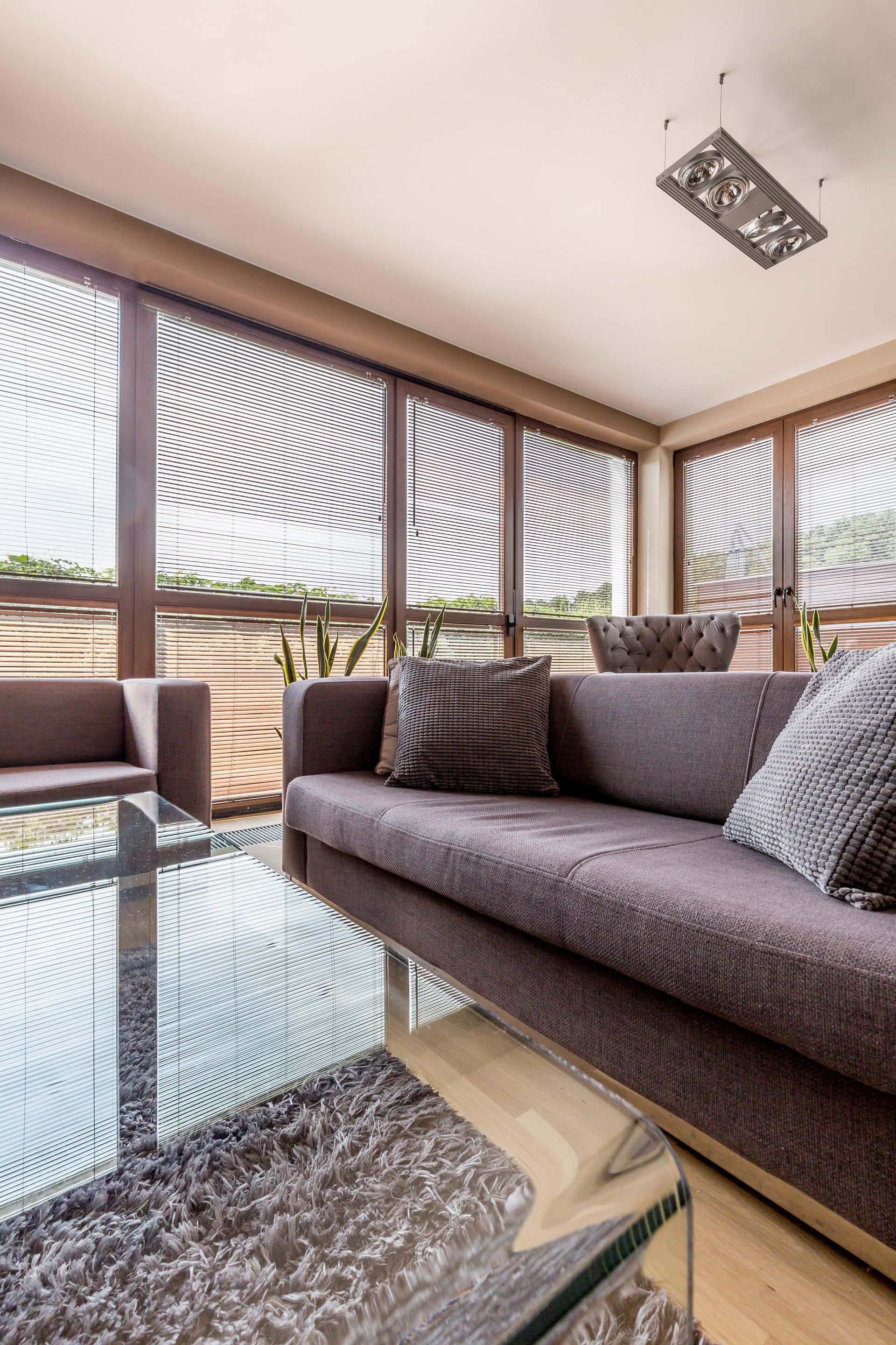 A modern living room with a dark grey sofa, glass coffee table, and large windows with decorative, curved wooden blinds.