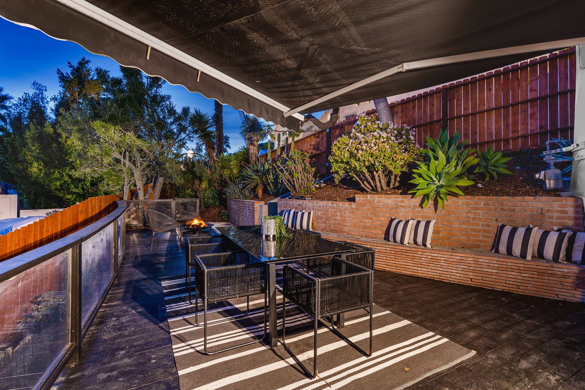 A deck dining area with a glass table, black chairs, and a striped rug under a retractable awning at dusk.