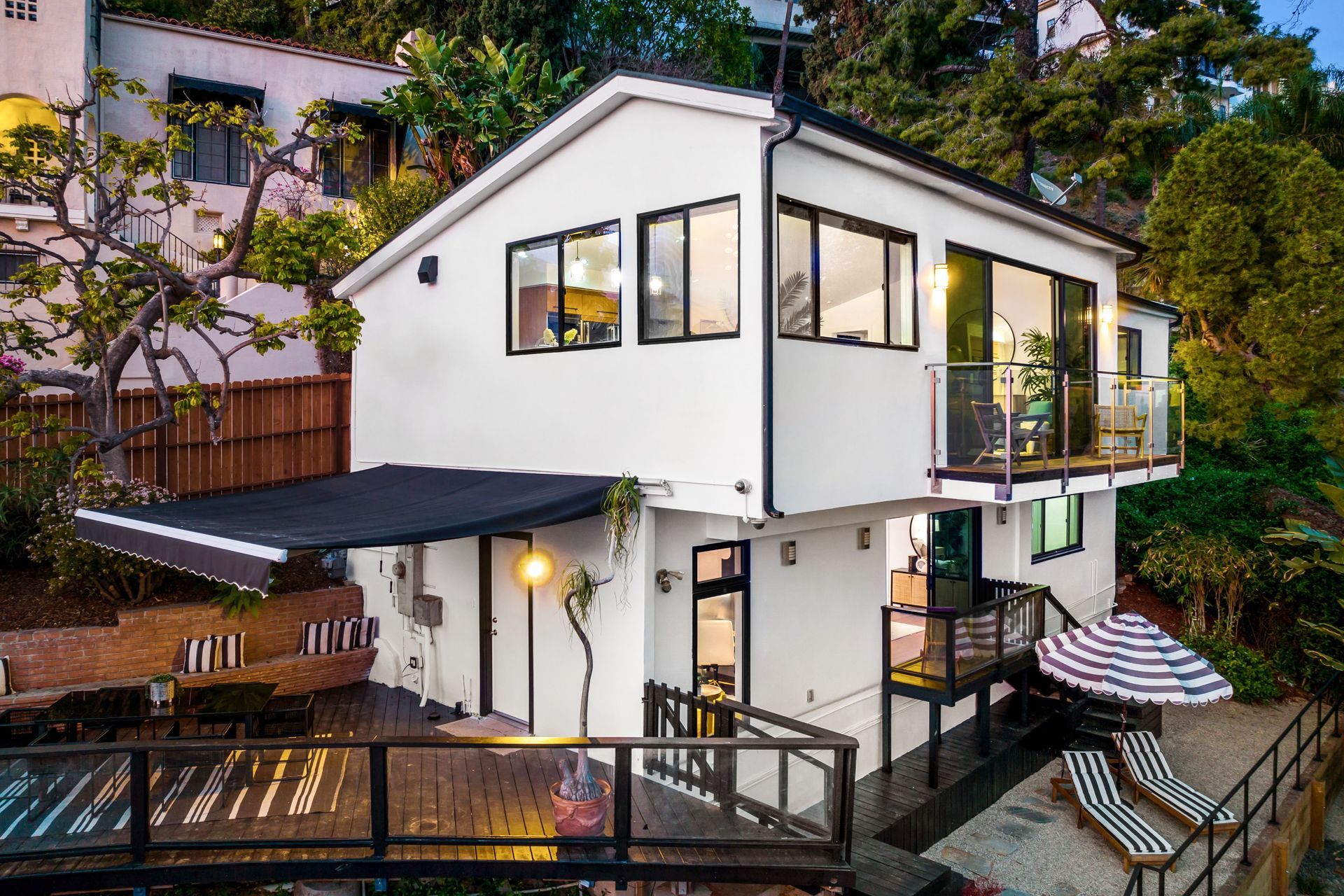 A two-story white house with a deck, black awning, and balcony, nestled on a hillside with a lower patio area.