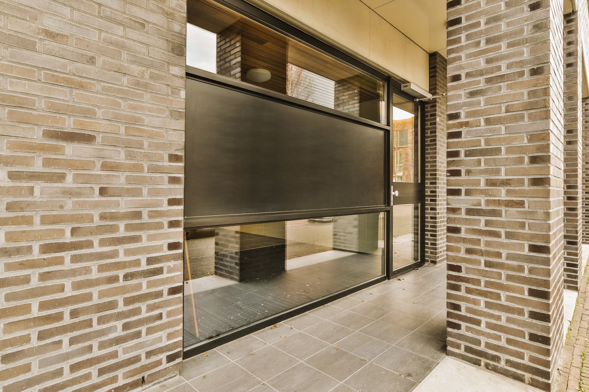 A modern exterior brick building facade featuring large glass windows partially covered by dark, retractable window shades.