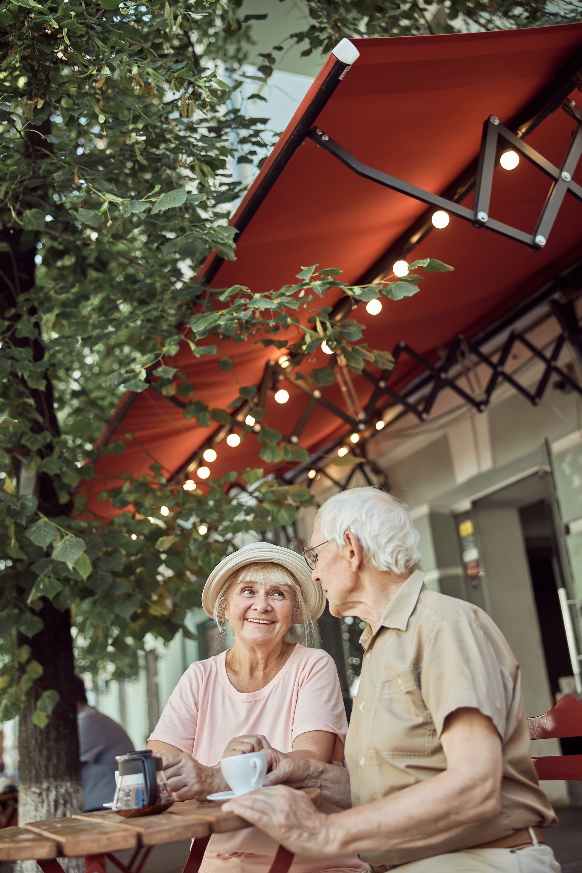 Two people sitting at an outdoor cafe table with drinks, smiling at each other under a red awning and string lights.