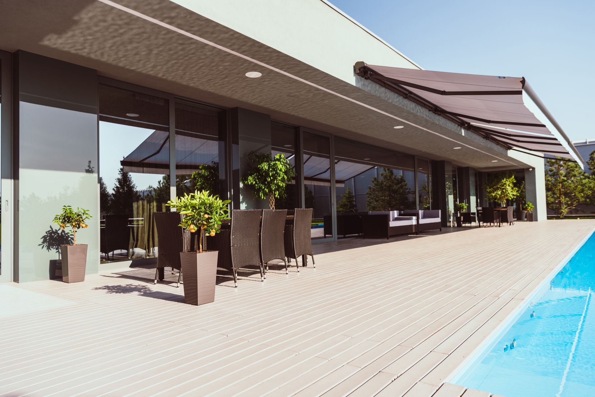 Modern outdoor patio with dining furniture and potted trees next to a swimming pool under a bright, clear sky.