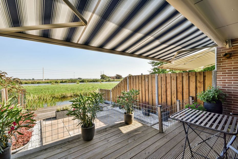 A wooden deck with potted plants and a small table under a striped awning, overlooking a grassy field and canal.