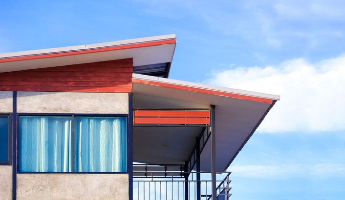 Modern house exterior featuring a flat, tiered roof, concrete walls, large windows with blue curtains, and a balcony.