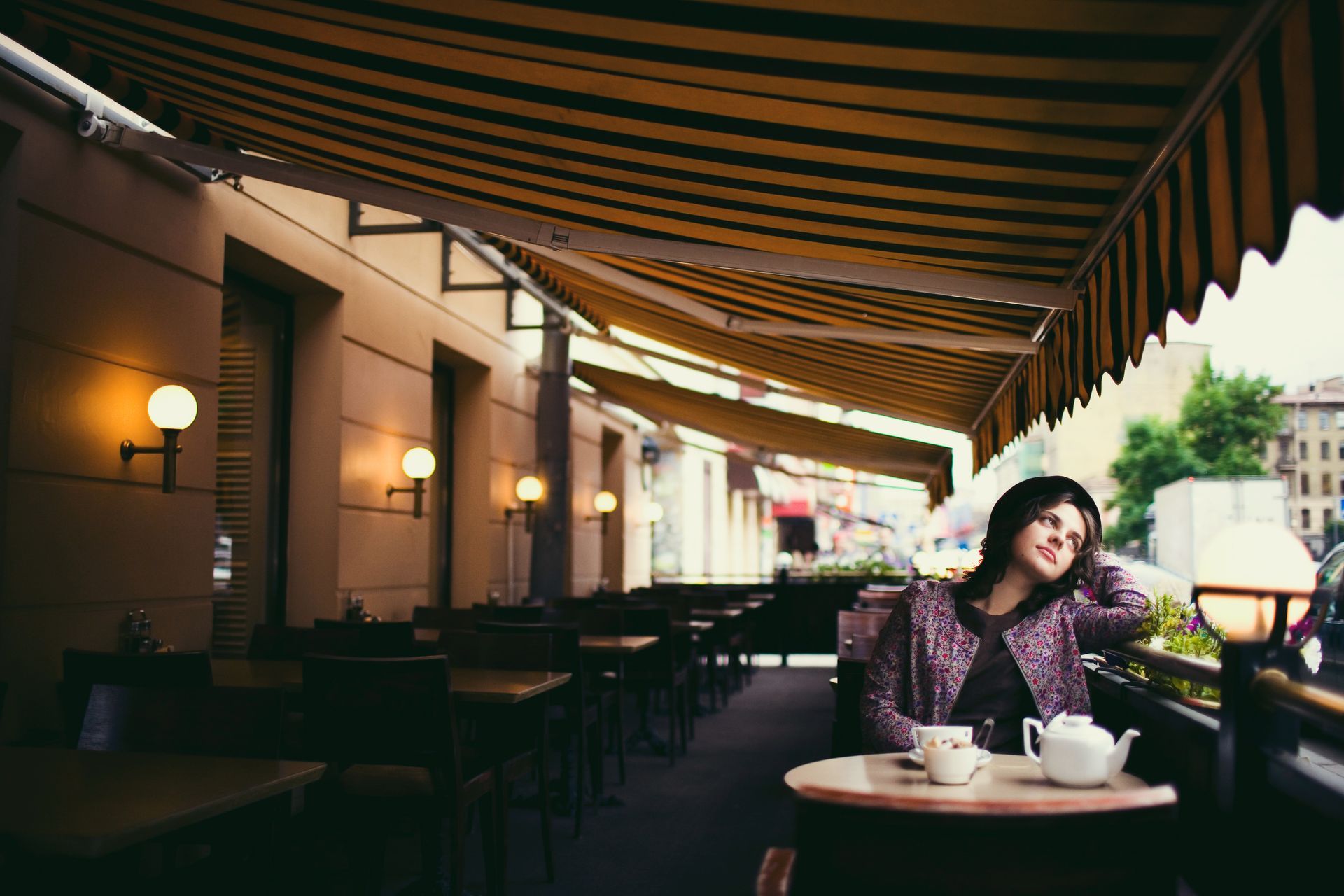A person sits at an outdoor cafe table under a striped awning, resting their head on their hand and looking thoughtfully.