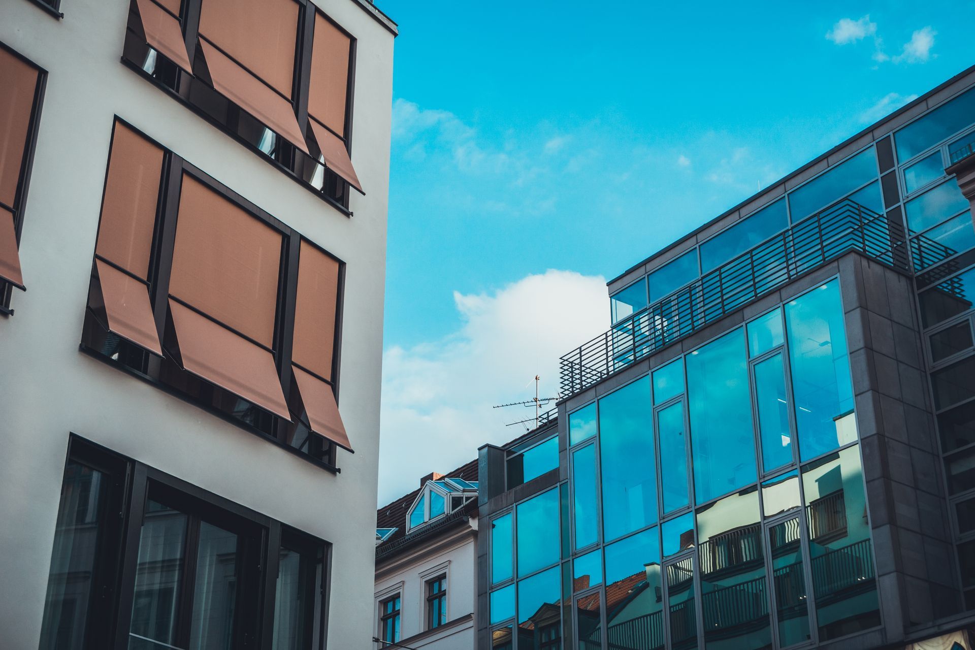 Two modern buildings with glass facades and brown window awnings stand under a bright blue, partly cloudy sky.
