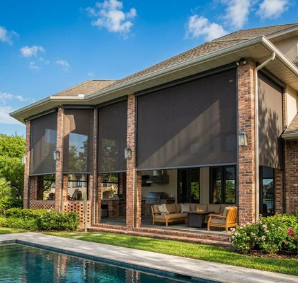 A spacious, covered outdoor patio featuring brick pillars, large solar screens, and comfortable seating by a swimming pool.