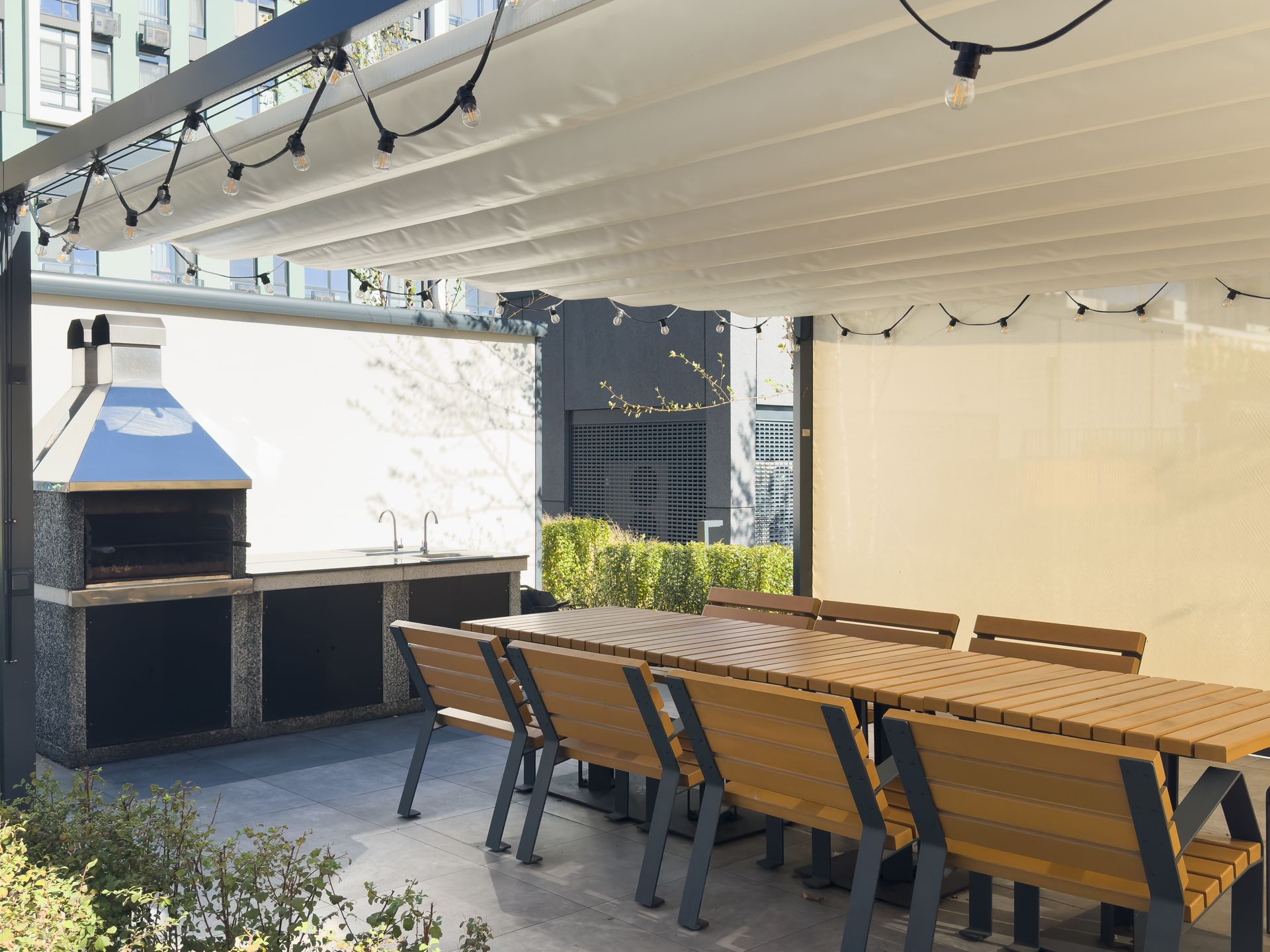 A patio with a large outdoor dining table, a built-in stone grill, and string lights under a retractable canopy.