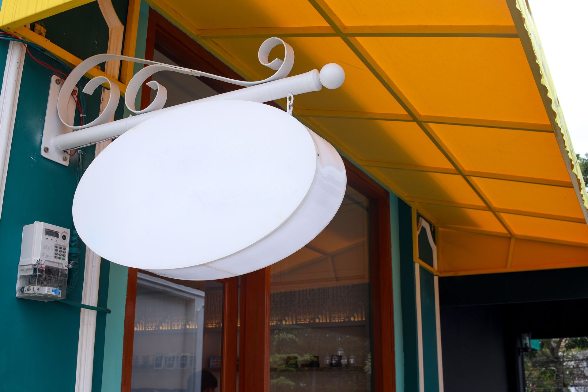 A blank, white oval sign hangs from a decorative white bracket against a teal wall, under a bright yellow fabric awning.