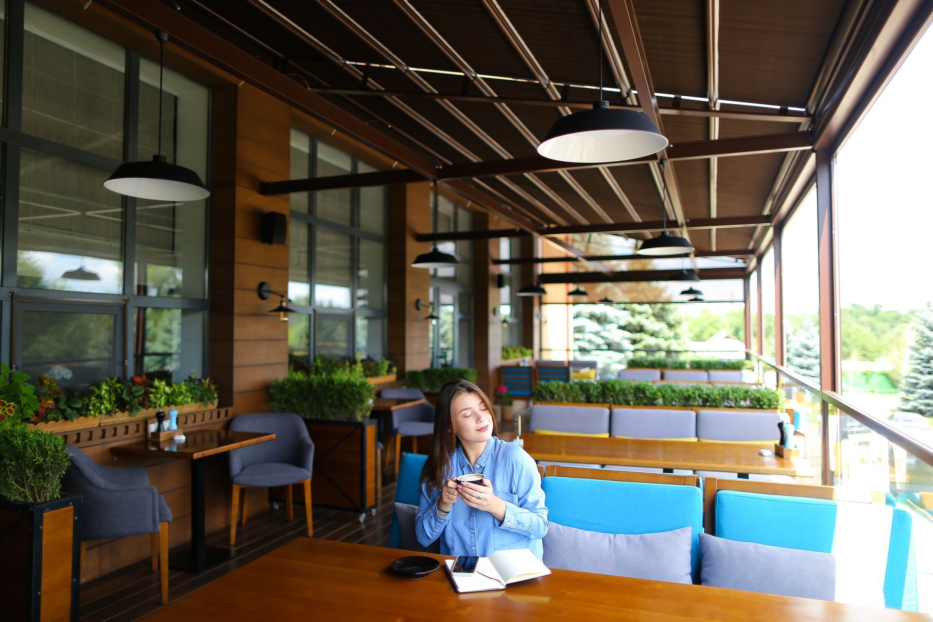 A person in a blue shirt sits at a wood table on an outdoor patio, holding a cup with a relaxed expression.