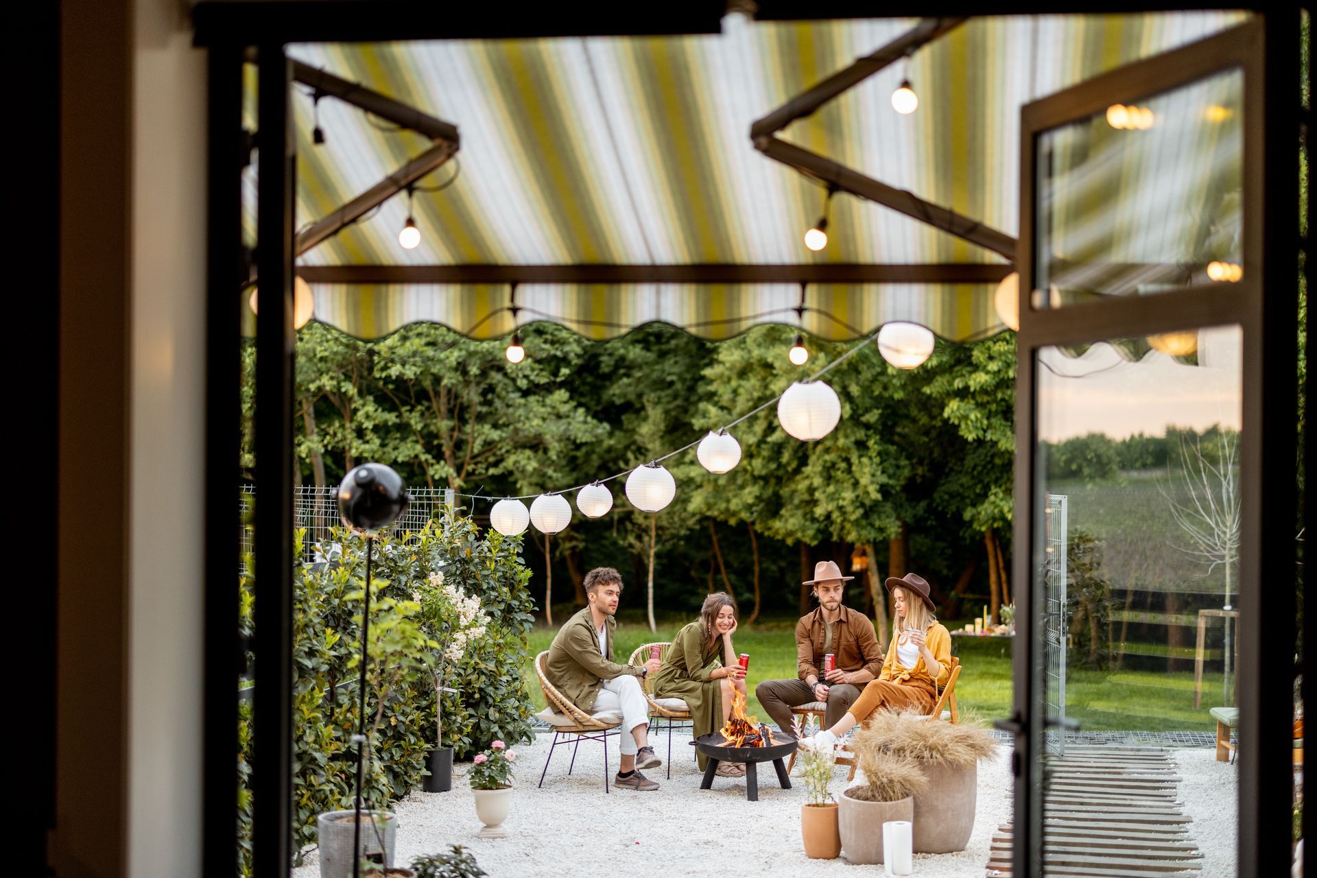 Three people sit in wicker chairs around a small fire pit on a patio under a striped awning with string lights.