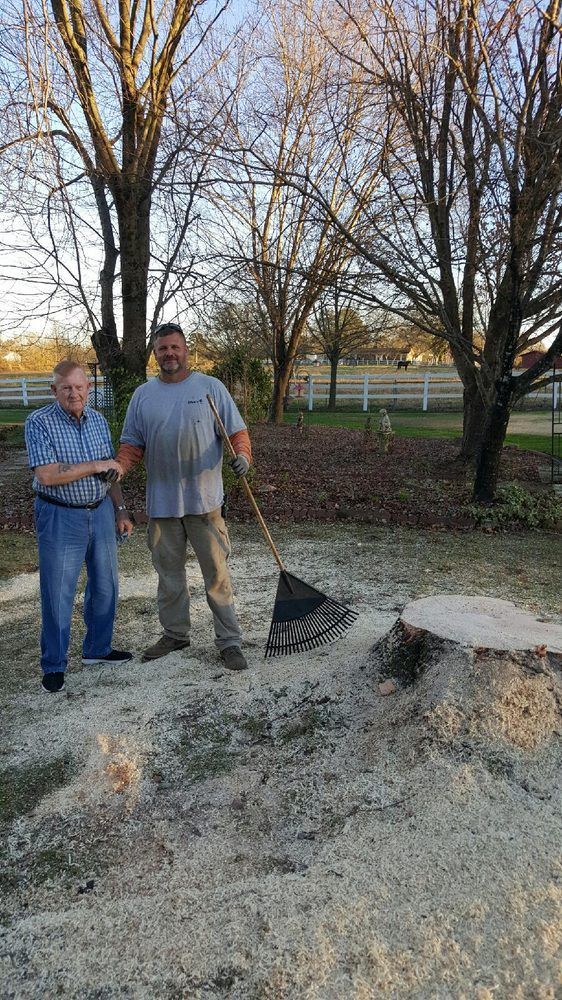 Two men are standing next to a tree stump with a rake.