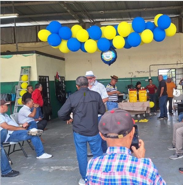 Gente reunida en una sala con globos azules y amarillos. Una celebración o evento está en marcha.