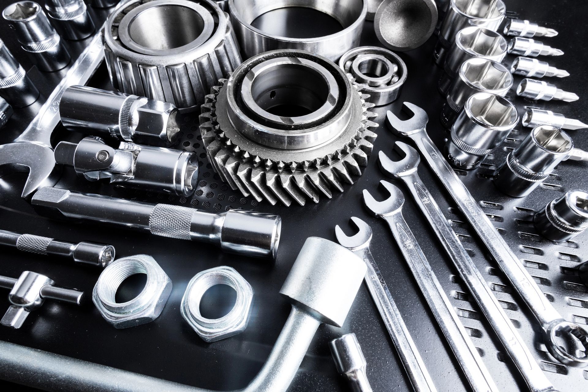 Primer plano de herramientas metálicas y piezas mecánicas, incluidas llaves, engranajes, cojinetes y enchufes sobre una superficie reflectante.