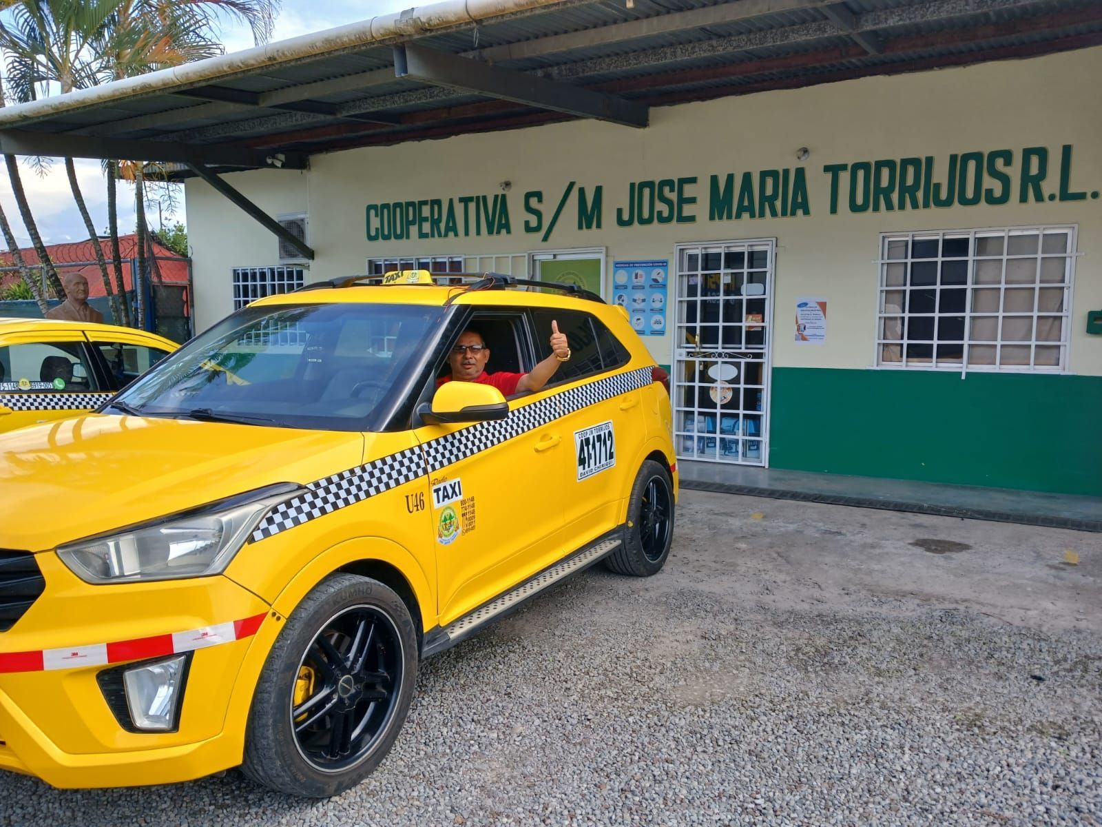 Taxi amarillo con conductor haciendo gesto de aprobación, estacionado frente a un edificio con el texto 