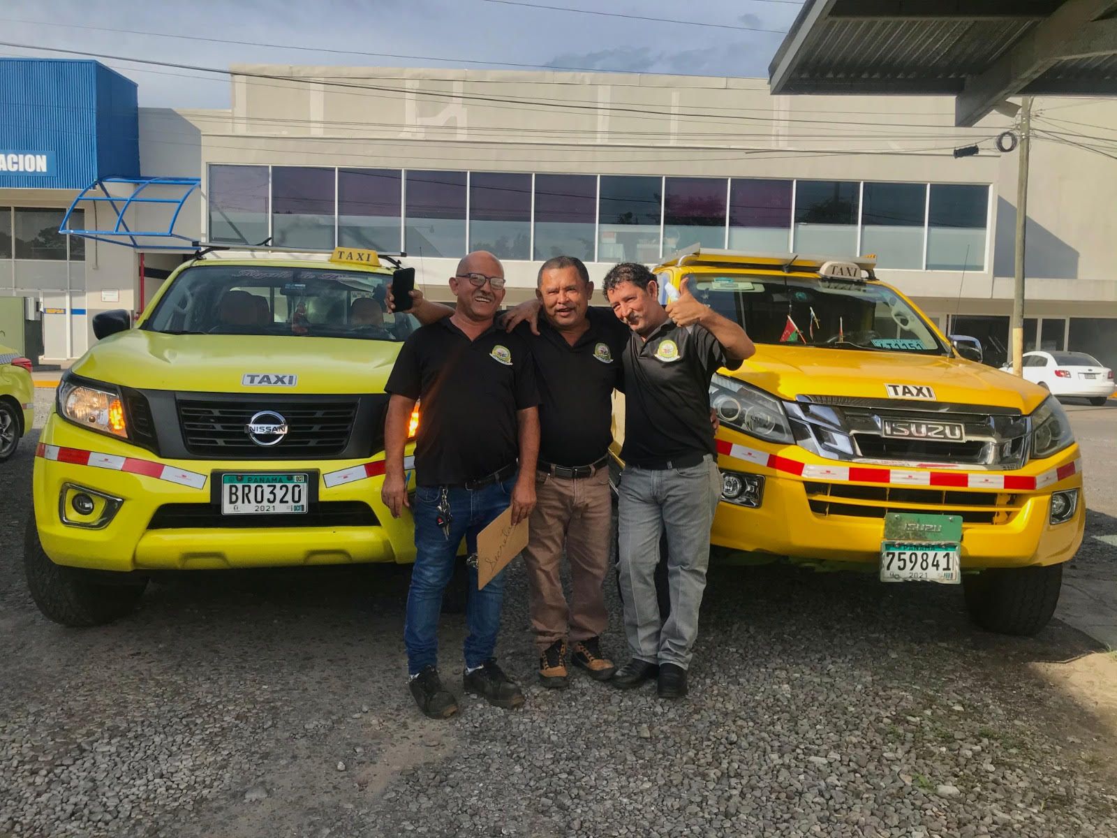 Tres hombres, sonrientes, están de pie junto a dos taxis amarillos. Los taxis están estacionados frente a un edificio.