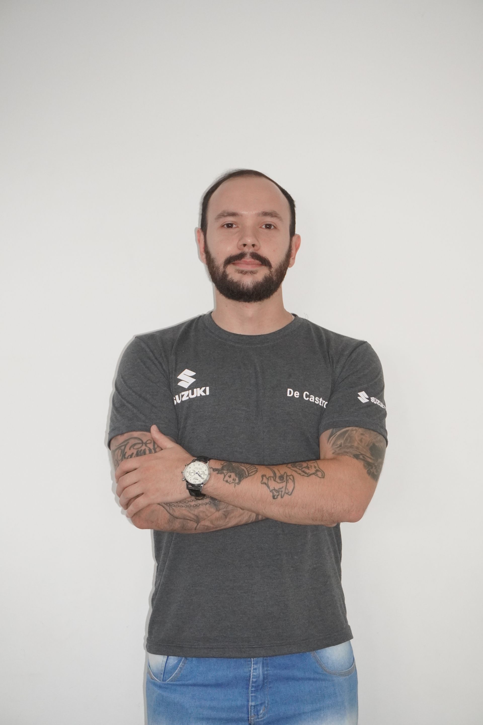 Homem barbudo e tatuado, vestindo uma camisa cinza, com os braços cruzados, em pé em frente a uma parede branca.