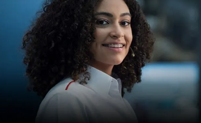 Mulher de cabelos cacheados sorri, vestindo uma camisa branca com detalhes em vermelho, olhando por cima do ombro.