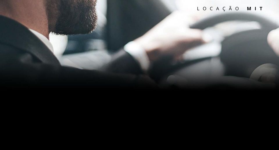 Homem de terno dirigindo um carro, mãos no volante. Interior desfocado, luz forte. Texto: