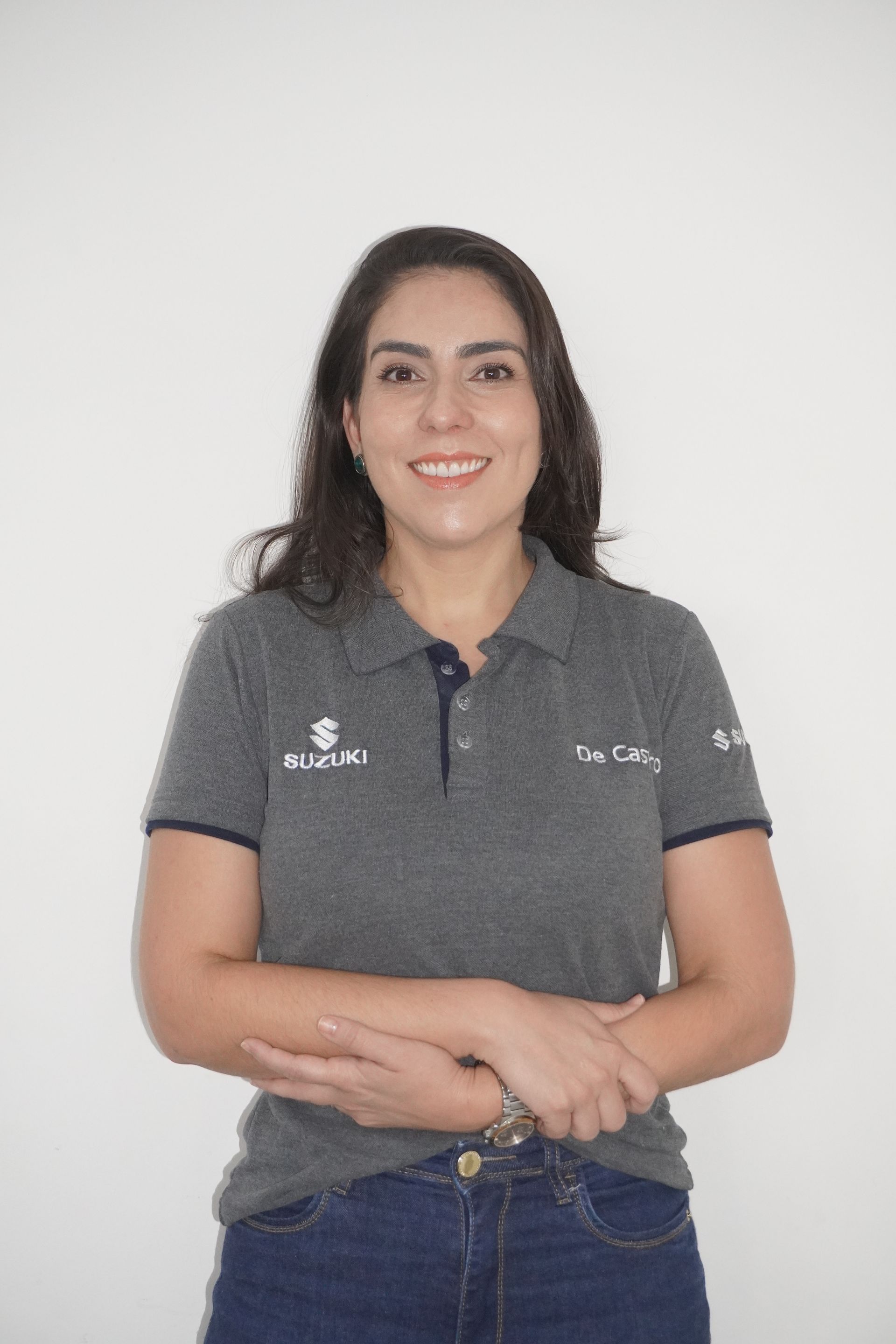 Mulher vestindo camisa polo cinza e calça jeans sorri, de braços cruzados, em frente a uma parede branca.