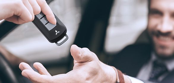 Pessoa entregando as chaves do carro para outra pessoa. Foco nas mãos e nas chaves; um homem sorridente está ao fundo.