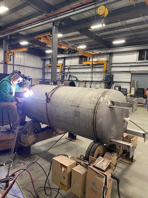 A man is welding a large stainless steel tank in a factory.