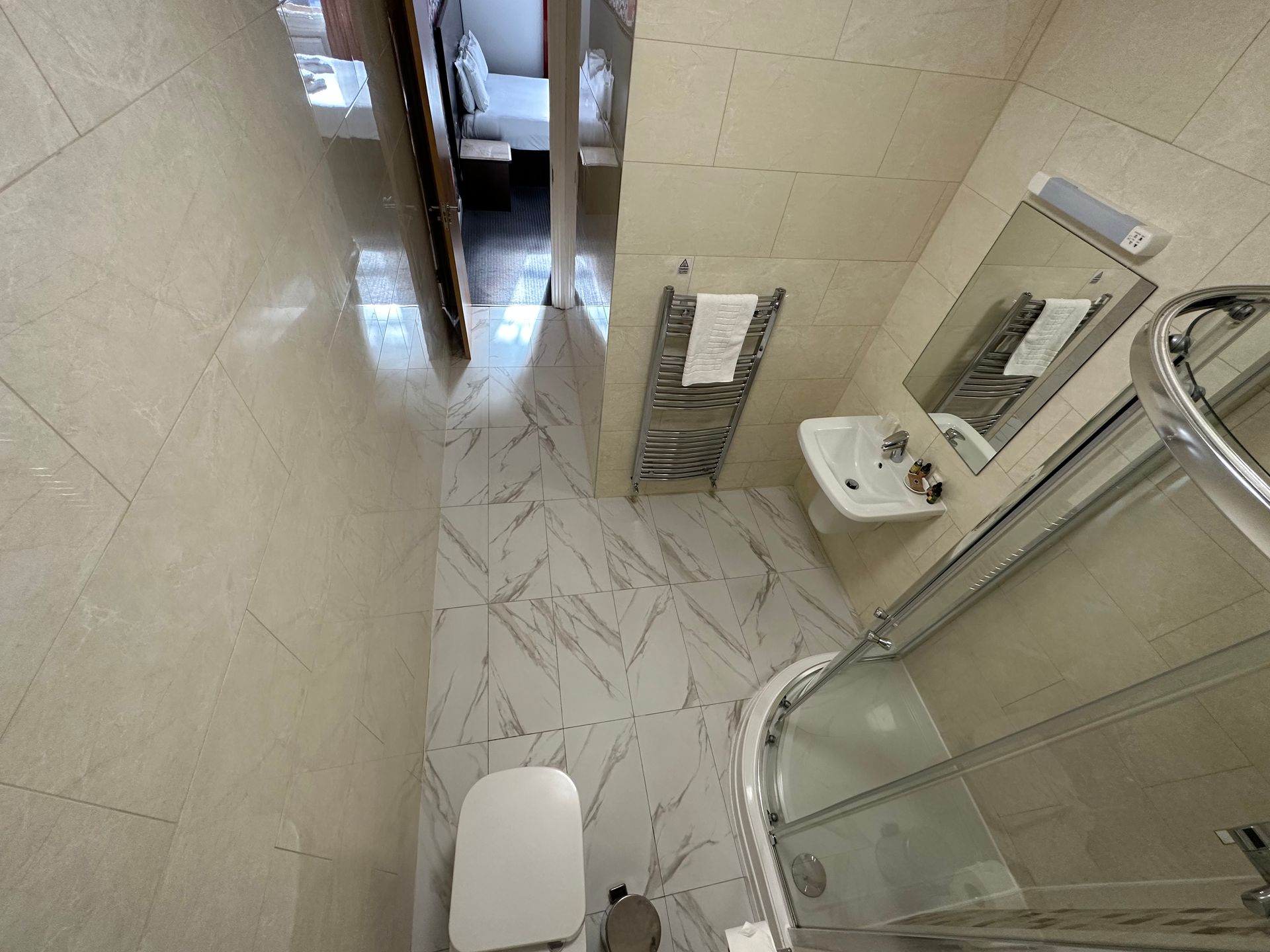Small bathroom with shower, toilet, sink, and towel rack. White and cream tile flooring and walls.