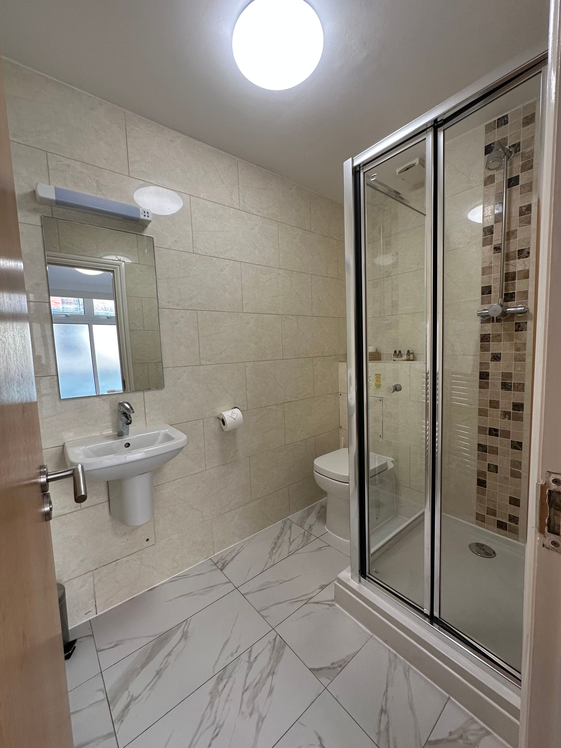 Bathroom with a shower, toilet, and sink. White and beige tiles, marble floors, and glass shower door.