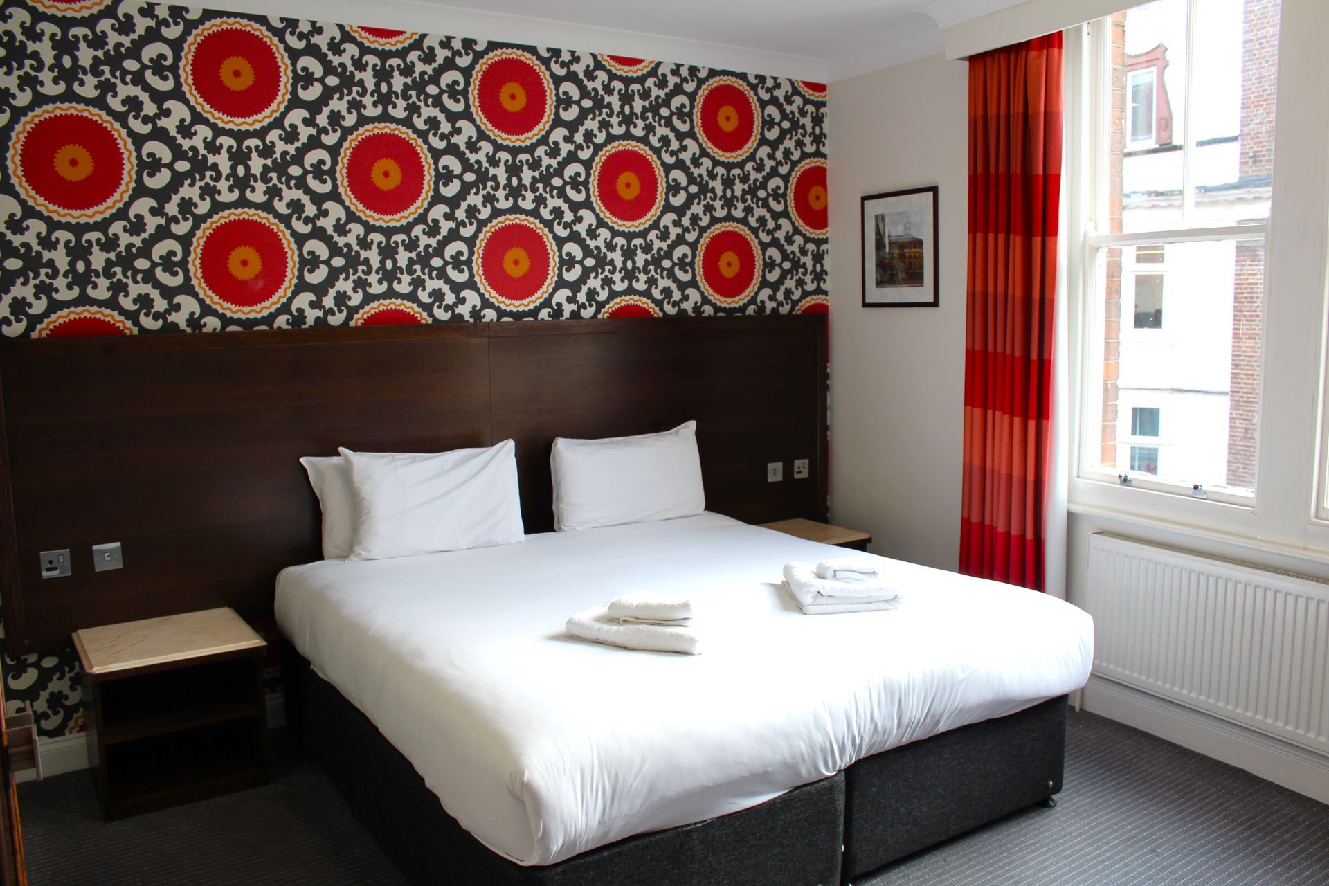 Bedroom with a patterned accent wall, bed, nightstands, red curtains, and a window.
