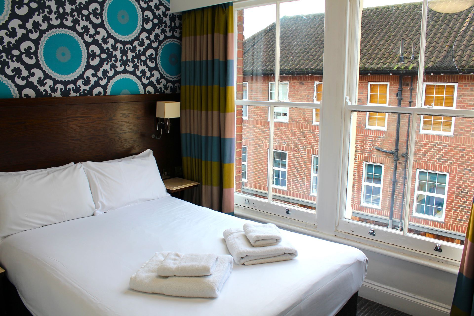Bedroom with a bed, towels, window, patterned wallpaper, and colorful curtains. Building visible outside the window.