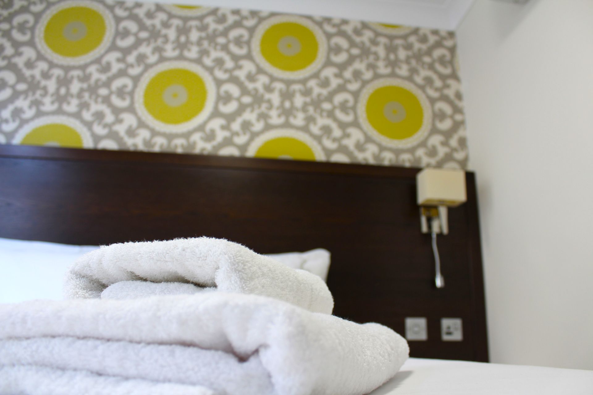 Folded white towels on a bed with a dark headboard, patterned wallpaper, and a wall lamp.
