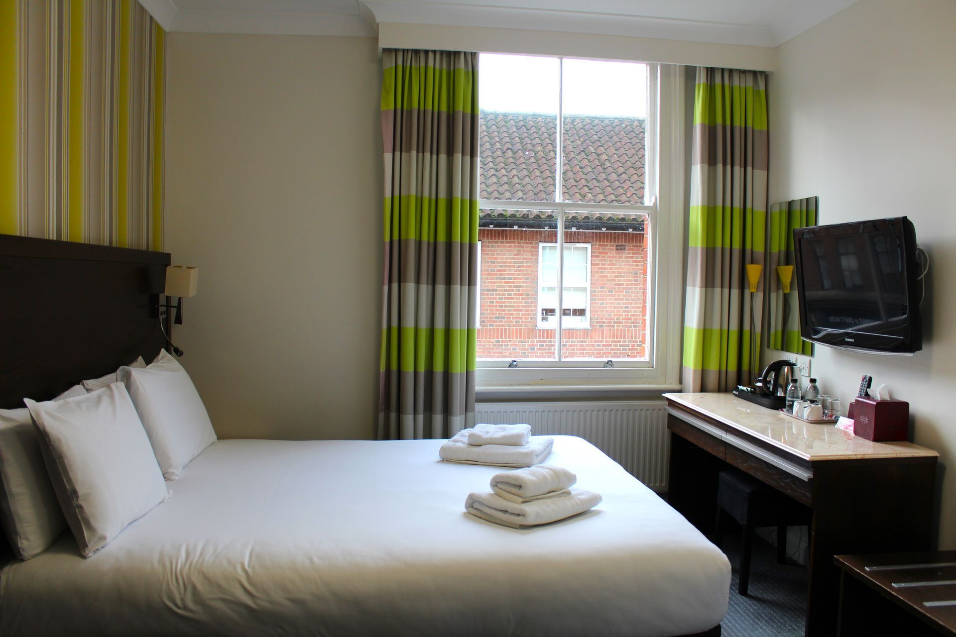 Hotel room with a bed, window, TV, and desk. Striped green and brown curtains.