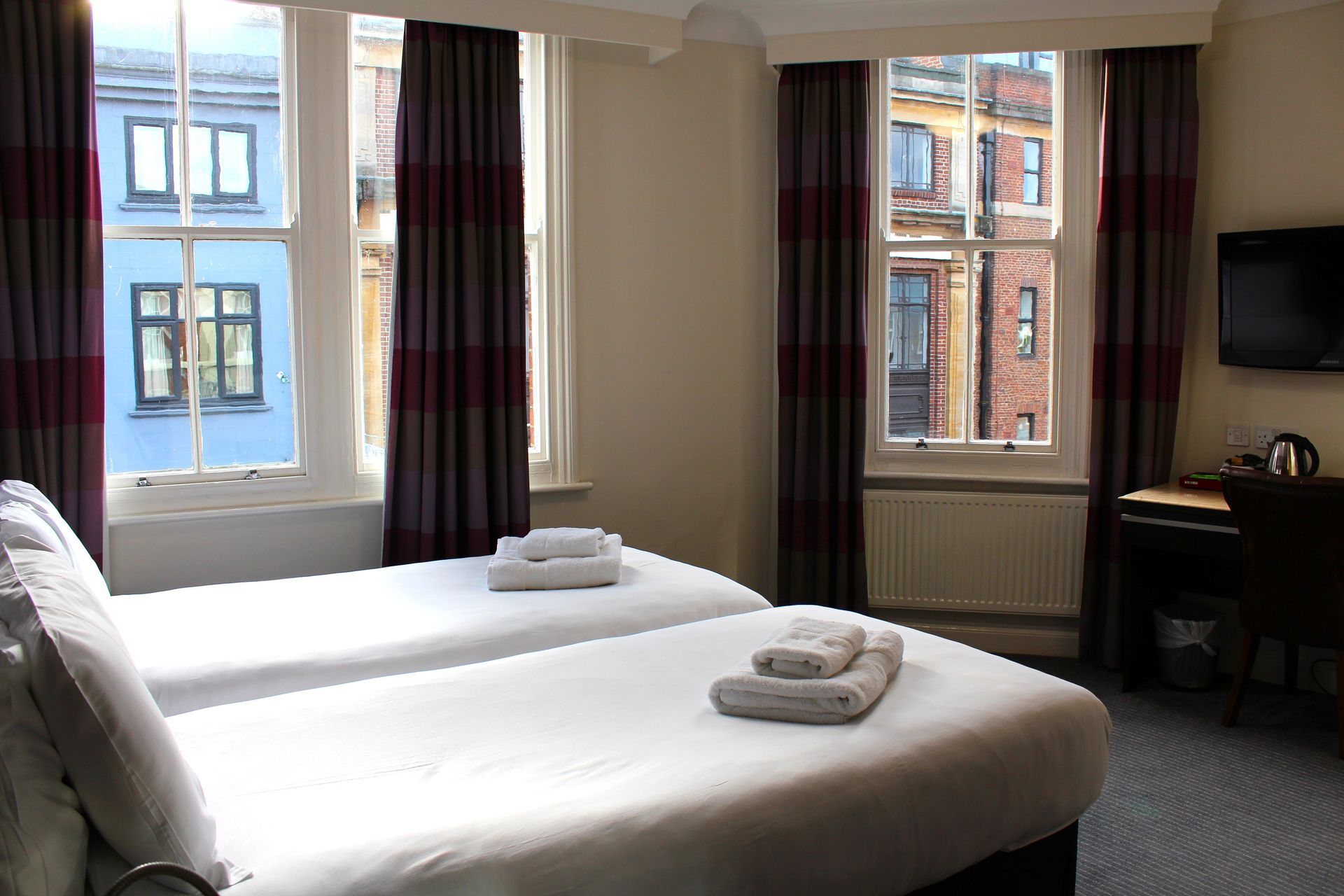Twin beds in a hotel room with windows, curtains, and a television.