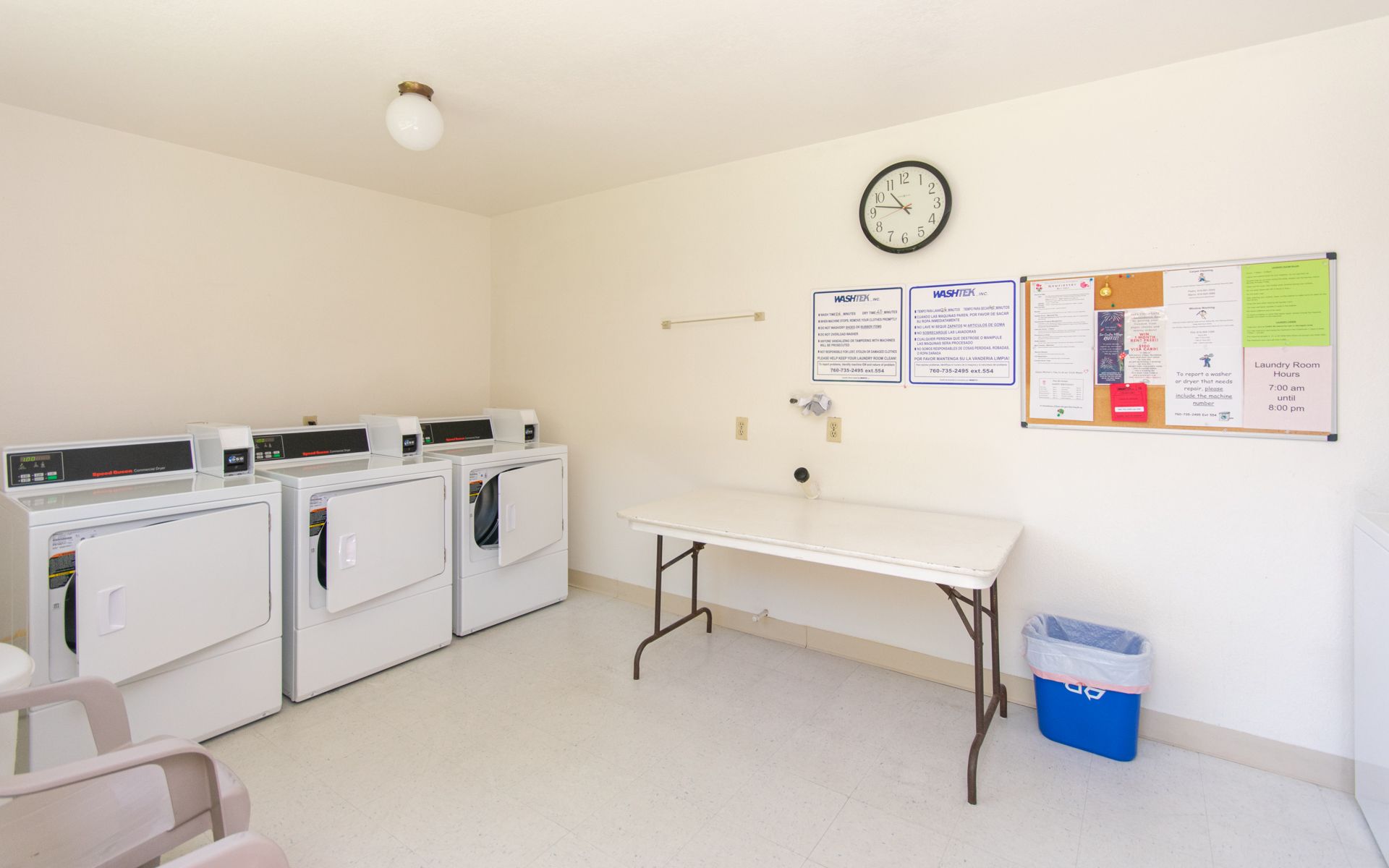 Laundry room with washers, dryer, folding table, bulletin board, clock, and trash can at Cowles Landing, offering apartments in San Diego, CA.
