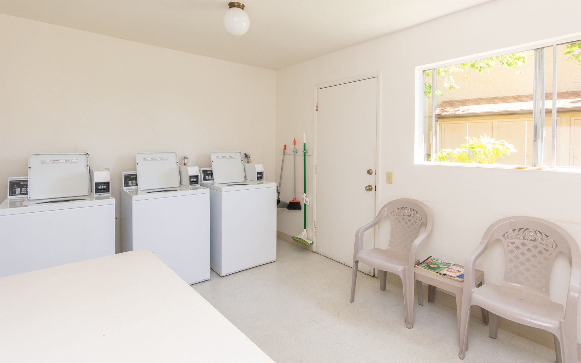 Laundry room with three washing machines, two chairs, and a window at Cowles Landing, offering apartments in San Diego, CA.