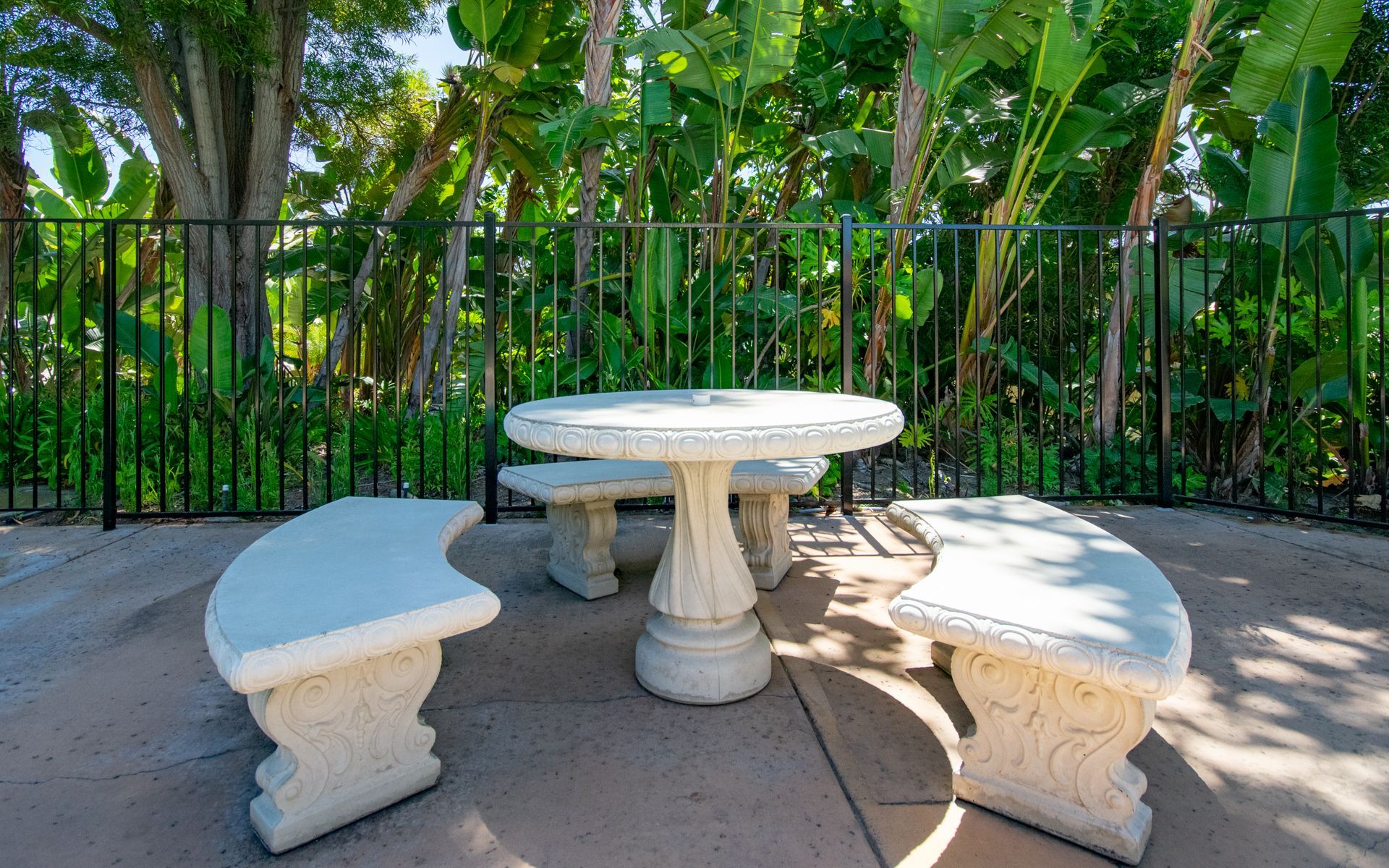 Stone table and benches in a garden, with a black fence and lush greenery in the background.