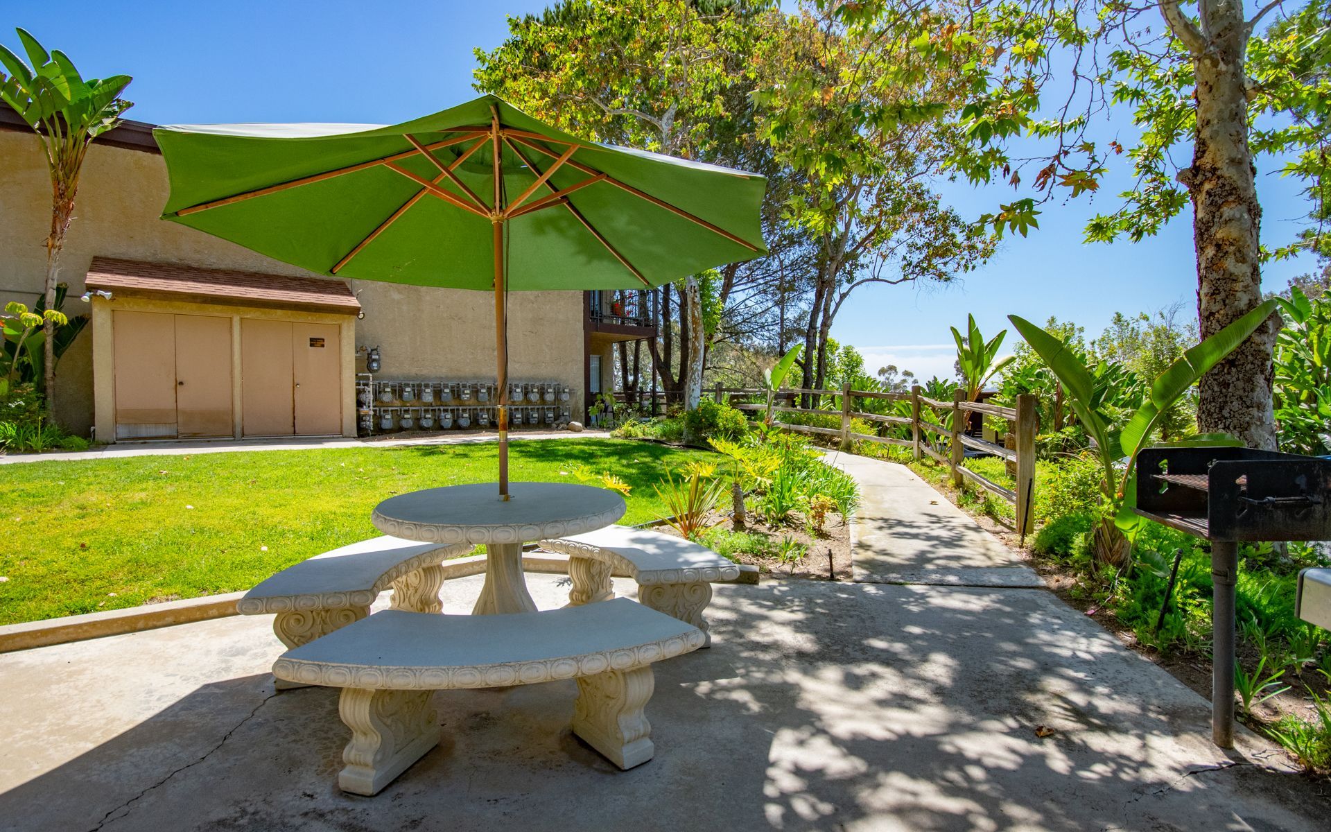 Picnic table with green umbrella on a paved walkway, lush greenery, and a building in the background.