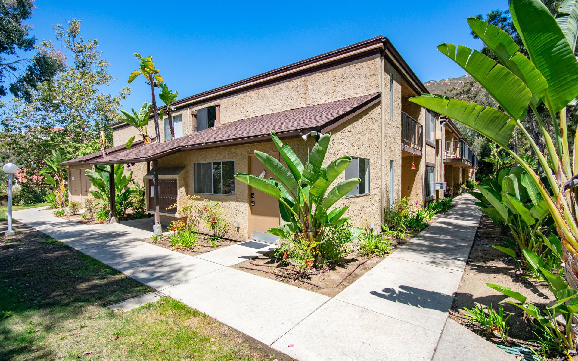 Two-story stucco apartment building with red-tiled roof, surrounded by tropical plants and a concrete walkway at Cowles Landing, offer apartments for rent in San Diego, CA.