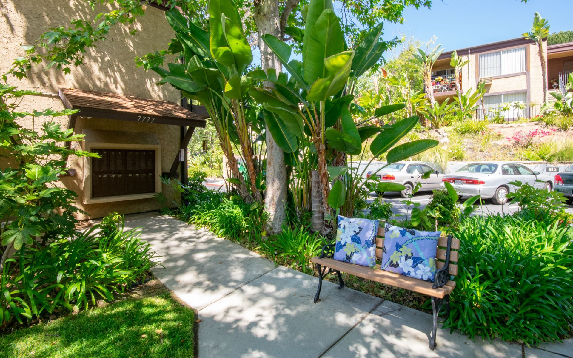 A bench with floral cushions sits on a sidewalk next to lush green foliage and a building at Cowles Landing, offering apartment living in San Diego, CA.