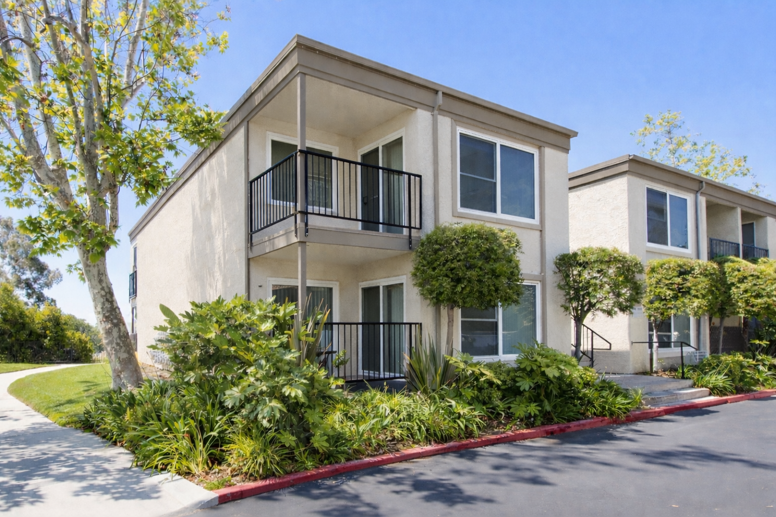 Two-story beige apartment building with balconies, shrubs, and sidewalk.